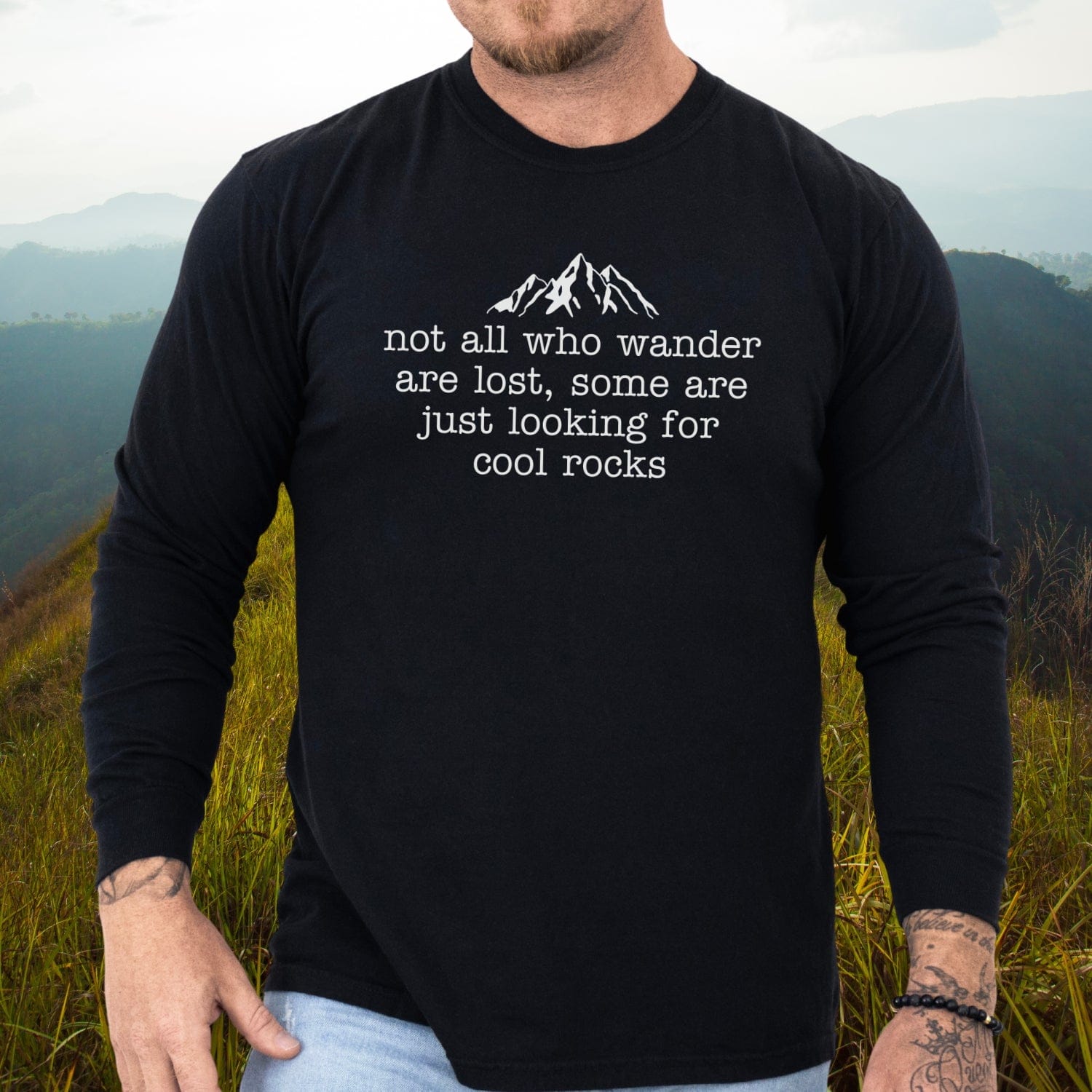 Person wearing Looking For Cool Rocks Comfort Colors Long-Sleeve Tee, showcasing mountain graphic and quote, standing on grassy hillside with visible forearm tattoo.