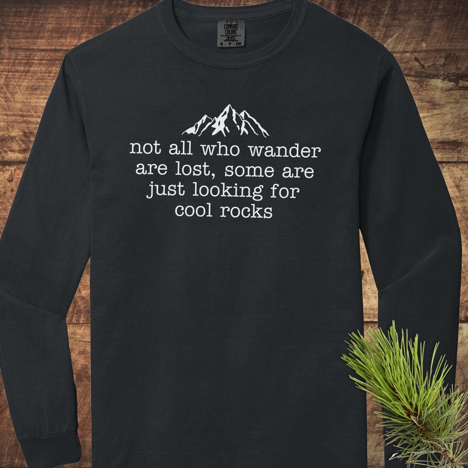 Looking For Cool Rocks Comfort Colors Long-Sleeve Tee features a mountain design and playful quote, laid out on a wooden surface with pine sprig.