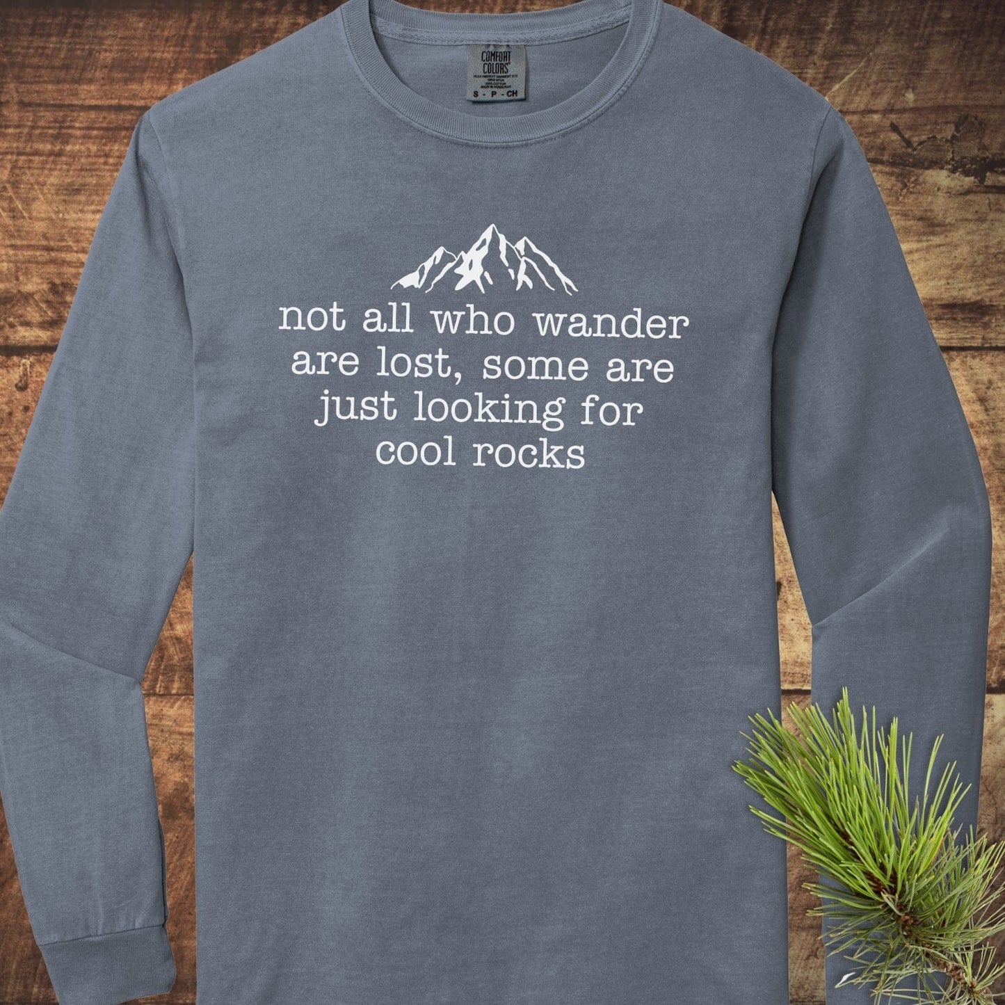 Looking For Cool Rocks Comfort Colors Long-Sleeve Tee, slate-blue, with mountain graphic and quote, laid on wood surface, next to pine sprig. 100% ring-spun cotton.
