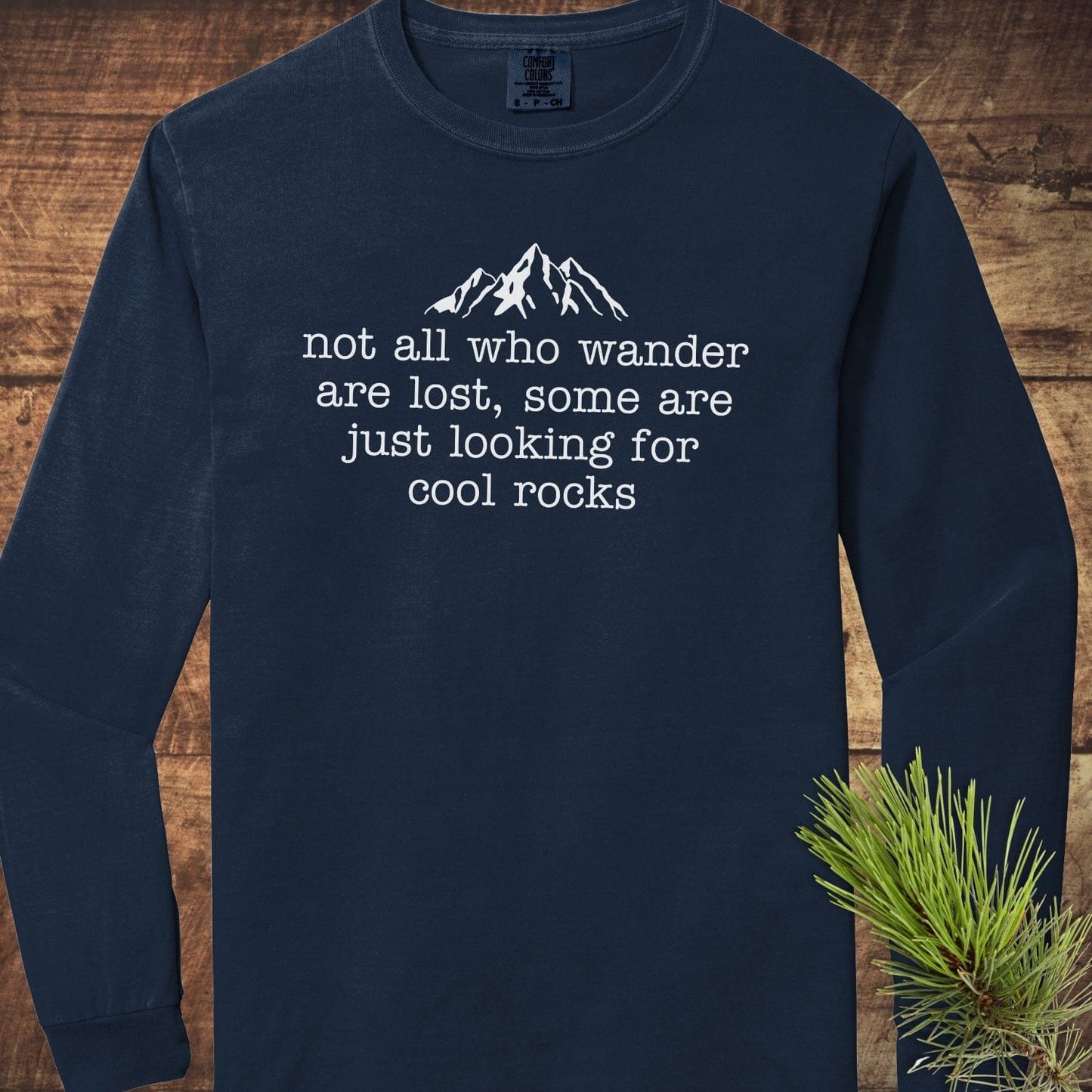 Looking For Cool Rocks Comfort Colors Long-Sleeve Tee featuring mountain peaks graphic and quote, laid flat with pine sprig for a rustic touch.