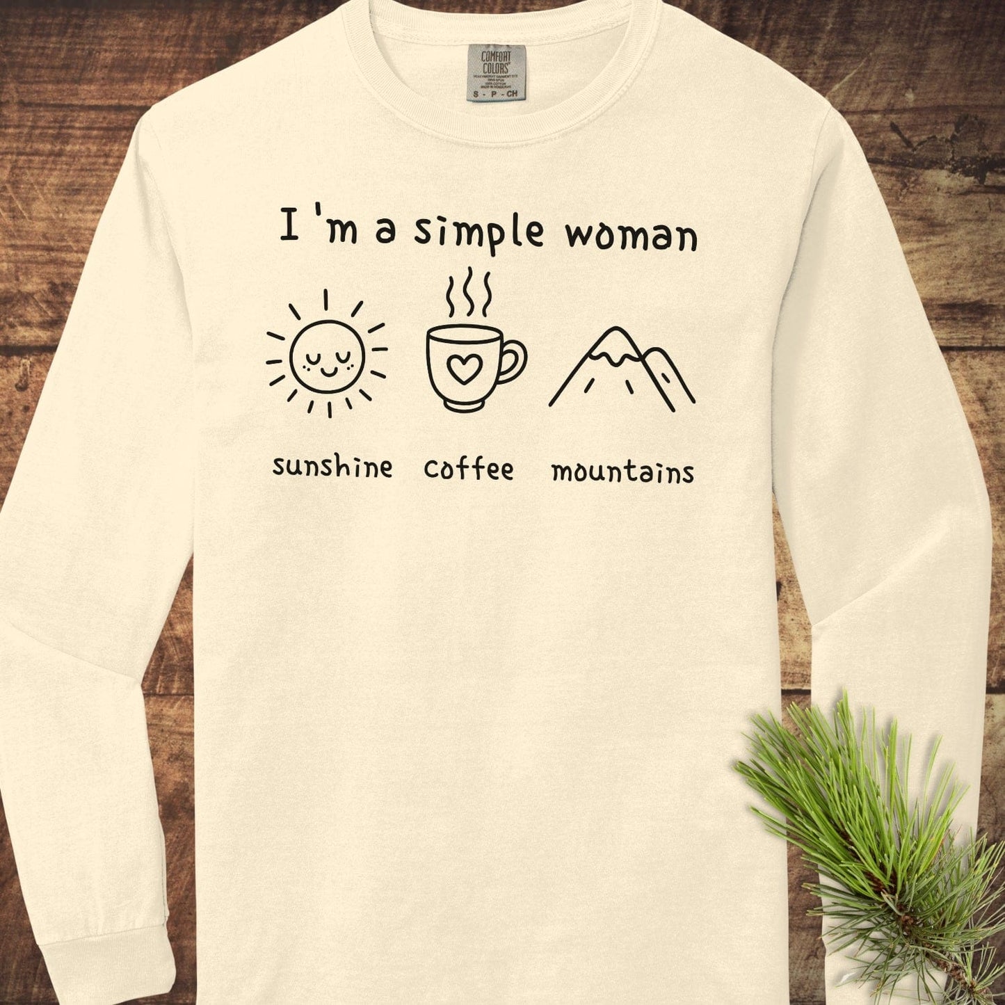 I'm A Simple Woman Comfort Colors Long-Sleeve Tee with sunshine, coffee, and mountains icons, laid flat with pine needle accent, showcasing soft, casual style.