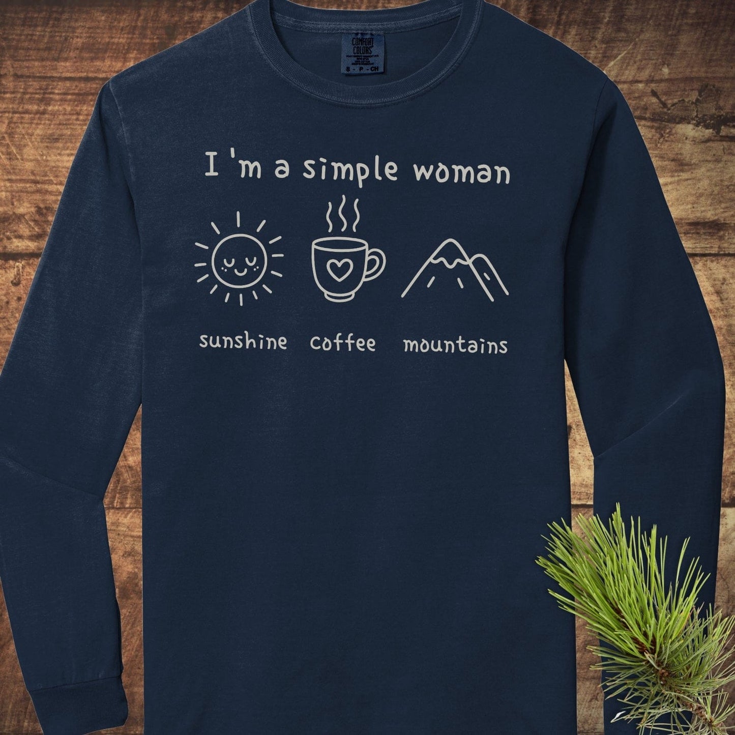 I'm A Simple Woman Comfort Colors Long-Sleeve Tee featuring 'sunshine,' 'coffee,' and 'mountains' doodles, displayed flat on wood, highlighting its relaxed fit and cotton softness.
