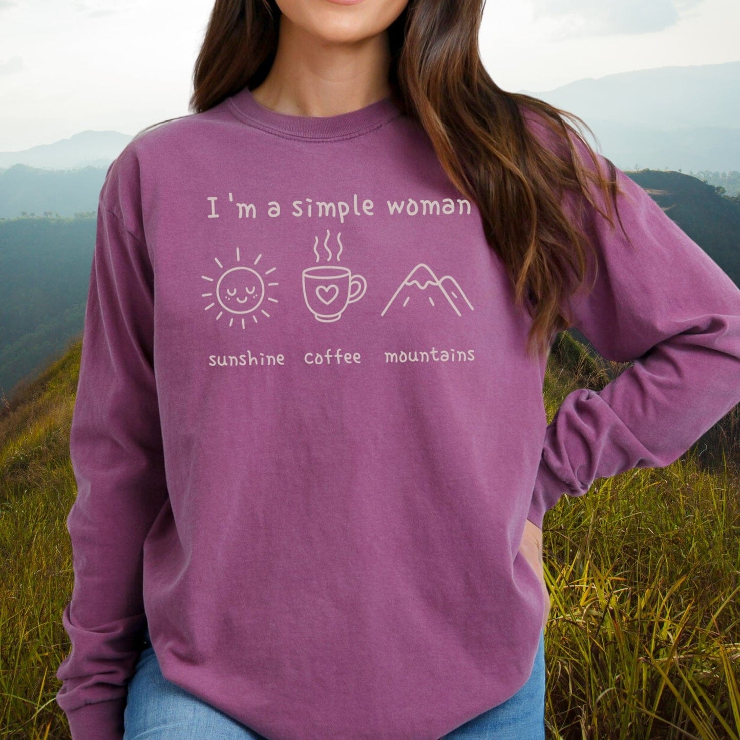 Woman wearing I'm A Simple Woman Comfort Colors Long-Sleeve Tee with sun, coffee, mountains icons, standing in a grassy, hilly landscape.