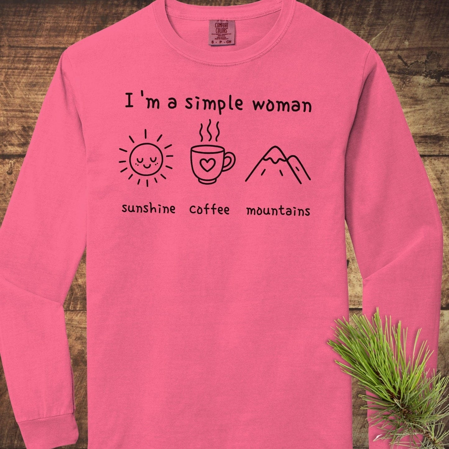 I'm A Simple Woman Comfort Colors Long-Sleeve Tee, pink, featuring sunshine, coffee, and mountains icons, laid on rustic wood with a pine sprig.