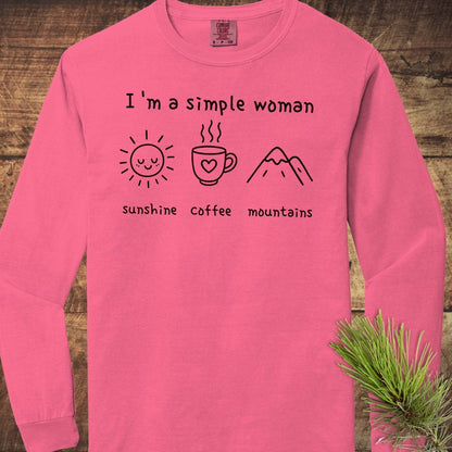 I'm A Simple Woman Comfort Colors Long-Sleeve Tee, pink, featuring sunshine, coffee, and mountains icons, laid on rustic wood with a pine sprig.