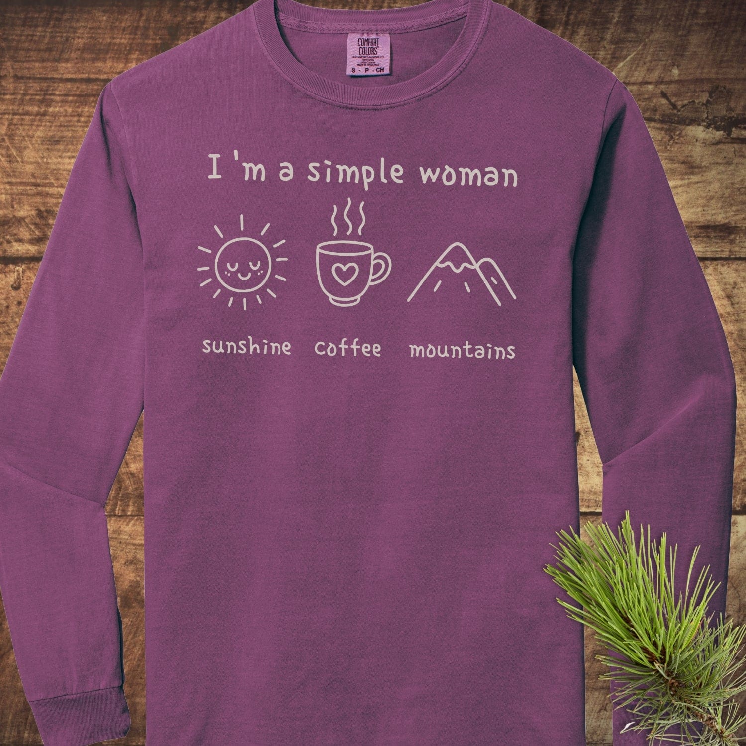 I'm A Simple Woman Comfort Colors Long-Sleeve Tee displayed on wood, featuring icons of a sun, coffee mug, and mountains, emphasizing casual comfort and style.