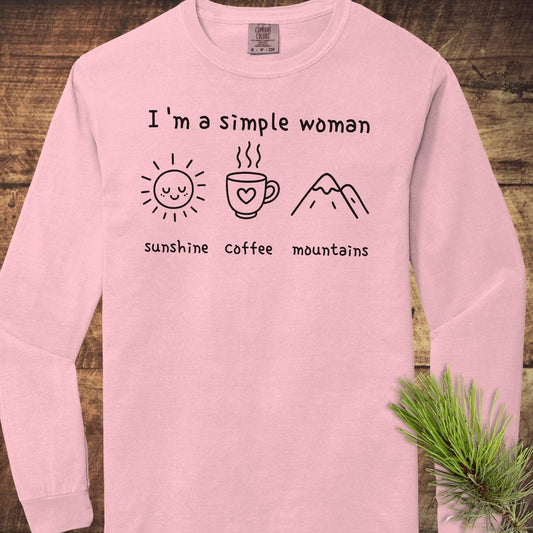 Light-pink I'm A Simple Woman long-sleeve tee on wood, featuring sun, coffee, mountains icons; soft, relaxed fit, 100% cotton.