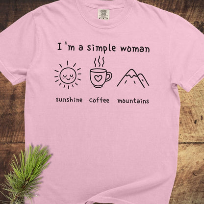 I'm A Simple Woman Comfort Colors 1717 Tee featuring icons of a sun, coffee mug, and mountains, on a wooden surface with a pine sprig.