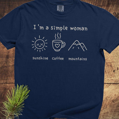 I'm A Simple Woman Comfort Colors 1717 Tee featuring a handwritten quote and icons of sunshine, coffee, and mountains, laid on a wooden surface.