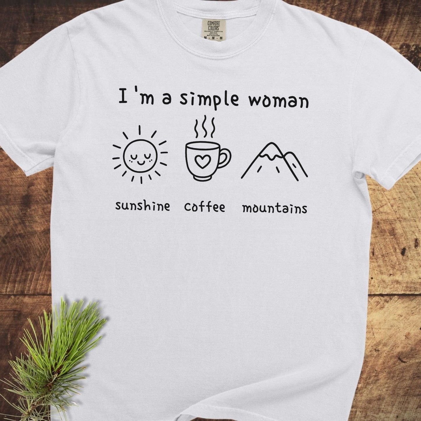 I'm A Simple Woman Comfort Colors 1717 Tee featuring minimalist line-art design with sunshine, coffee, and mountains icons, laid flat on a wooden surface.