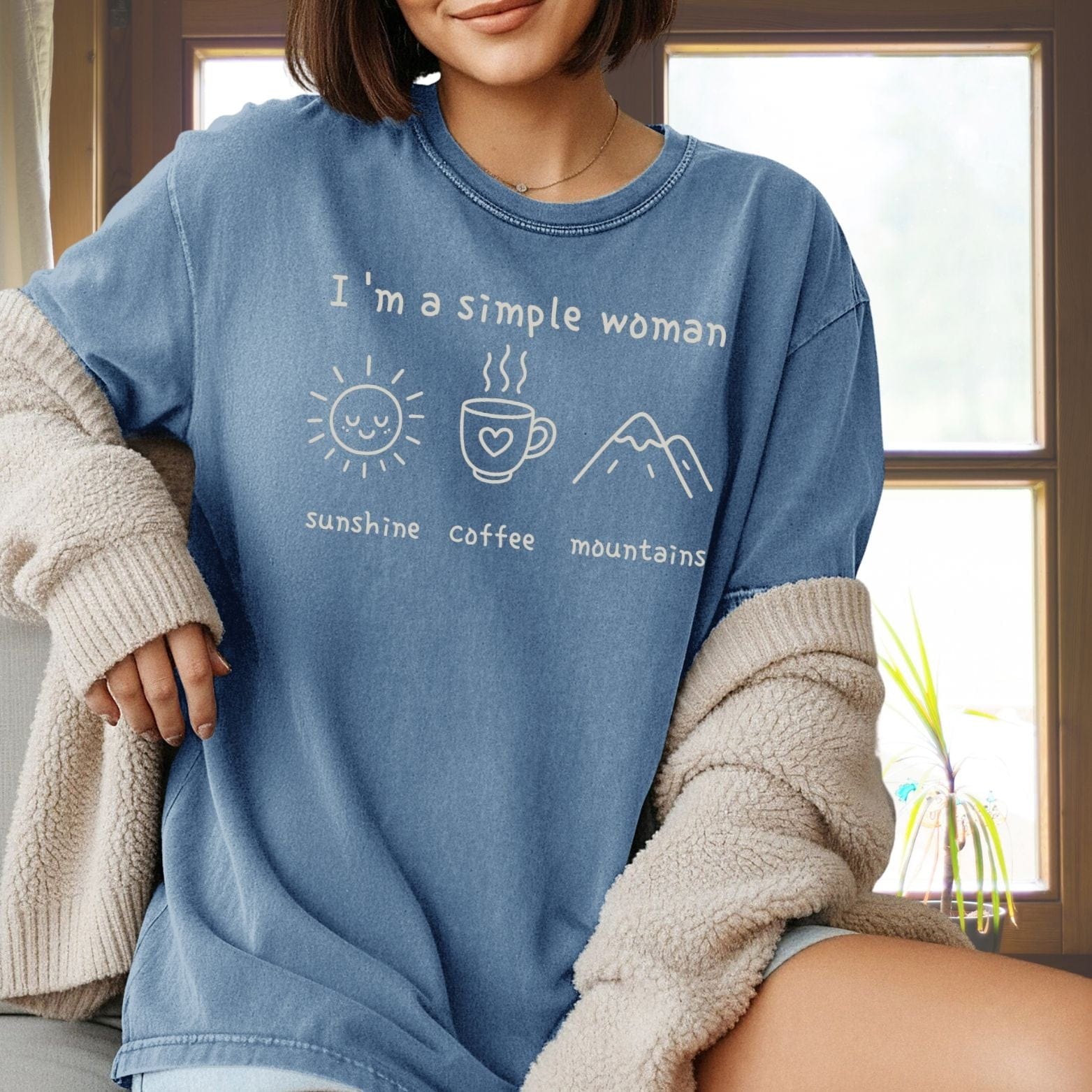 Woman wears I'm A Simple Woman Comfort Colors 1717 Tee, featuring sun, coffee, mountains icons, under a sherpa-style cardigan, sitting by a window.