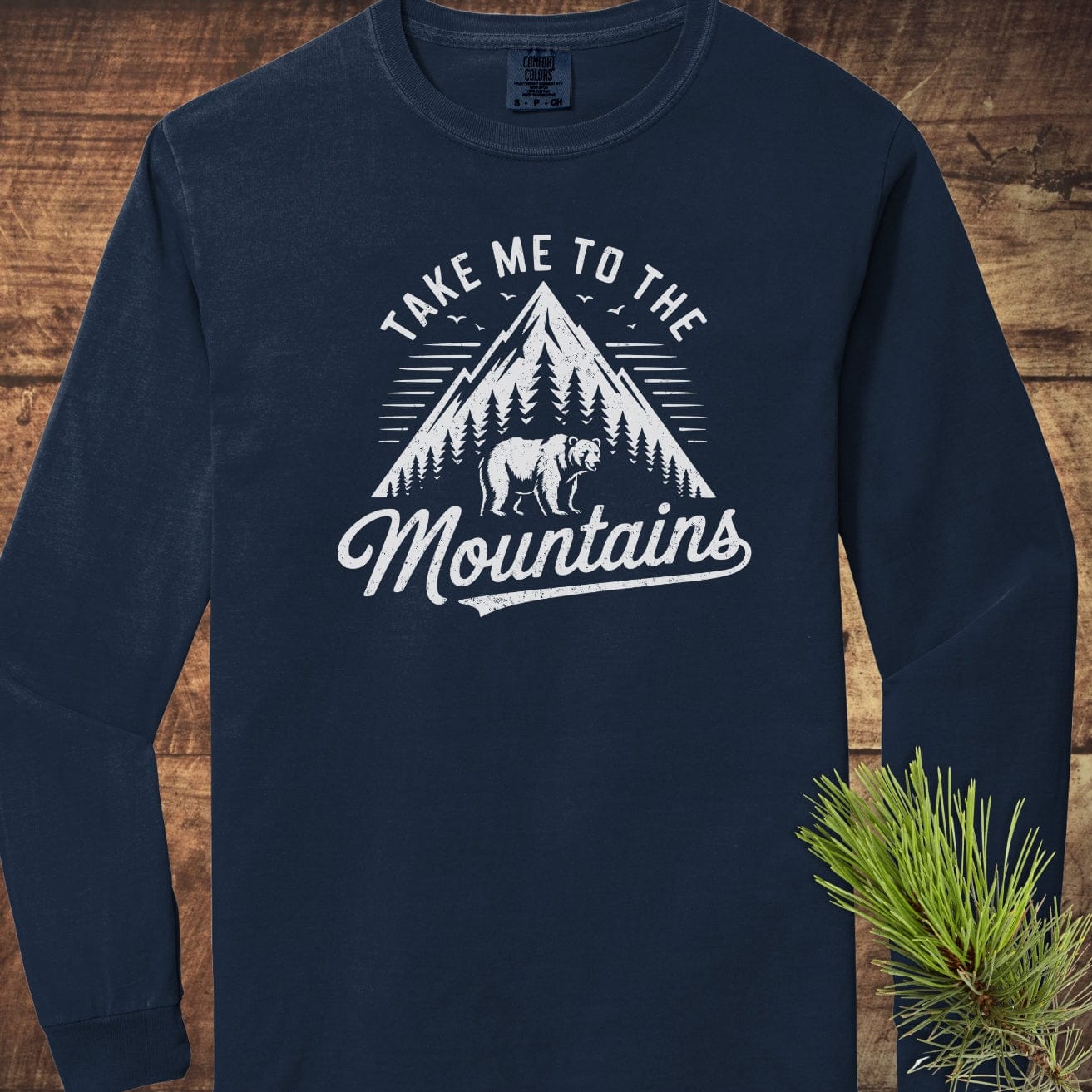 Copy of Mountain Energy Comfort Colors Long-Sleeve Tee featuring a mountain design and slogan, laid flat on a wooden surface with pine needles.