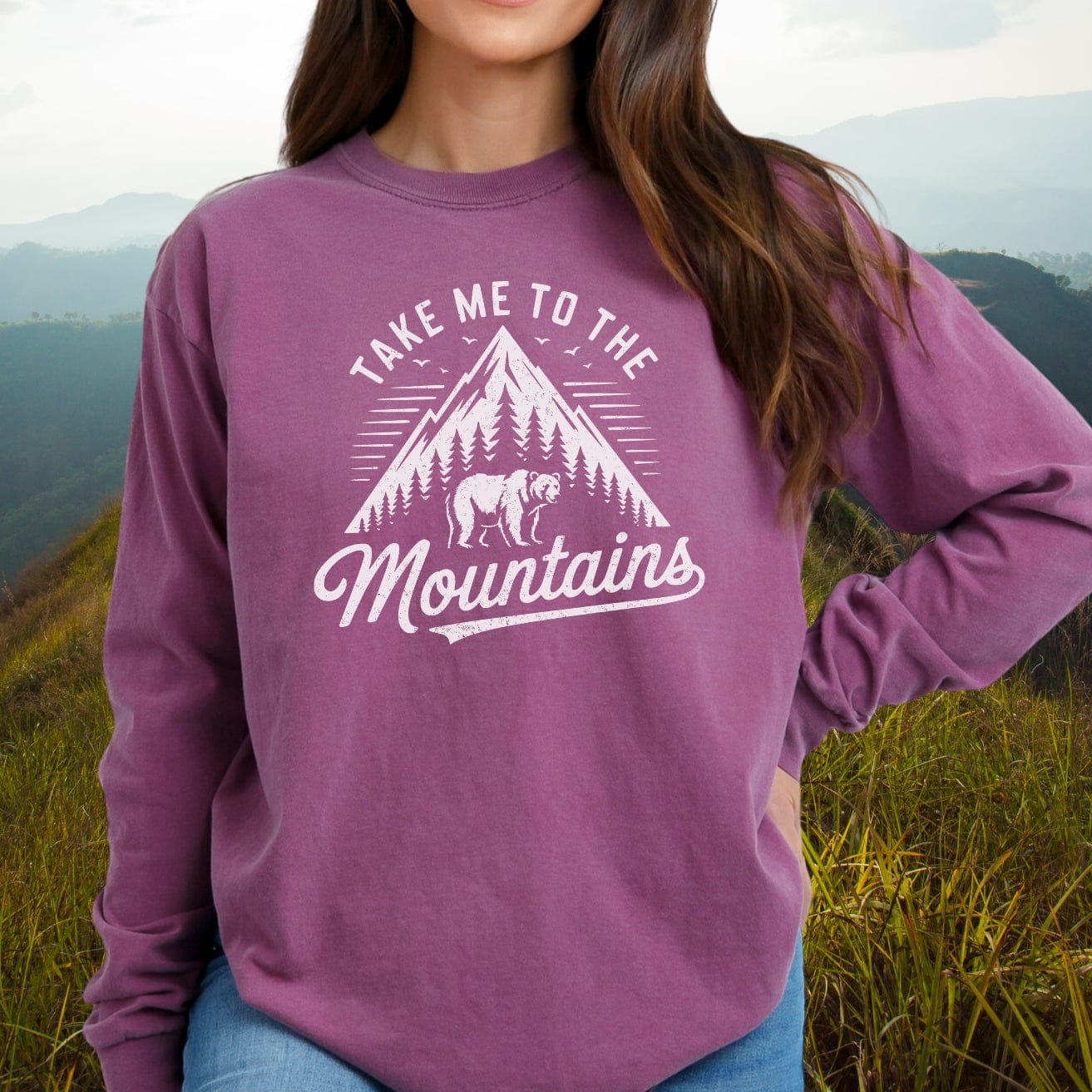 Person wearing the Copy of Mountain Energy Comfort Colors Long-Sleeve Tee, showcasing a mountain design, standing against grassy hills.