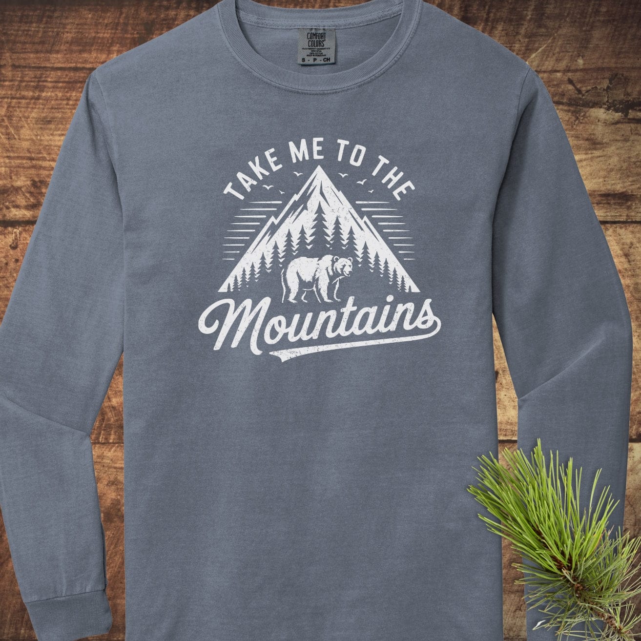 Copy of Mountain Energy Comfort Colors Long-Sleeve Tee featuring a mountain graphic, pine trees, bear silhouette, and “Take Me to the Mountains” slogan on rustic wood background.