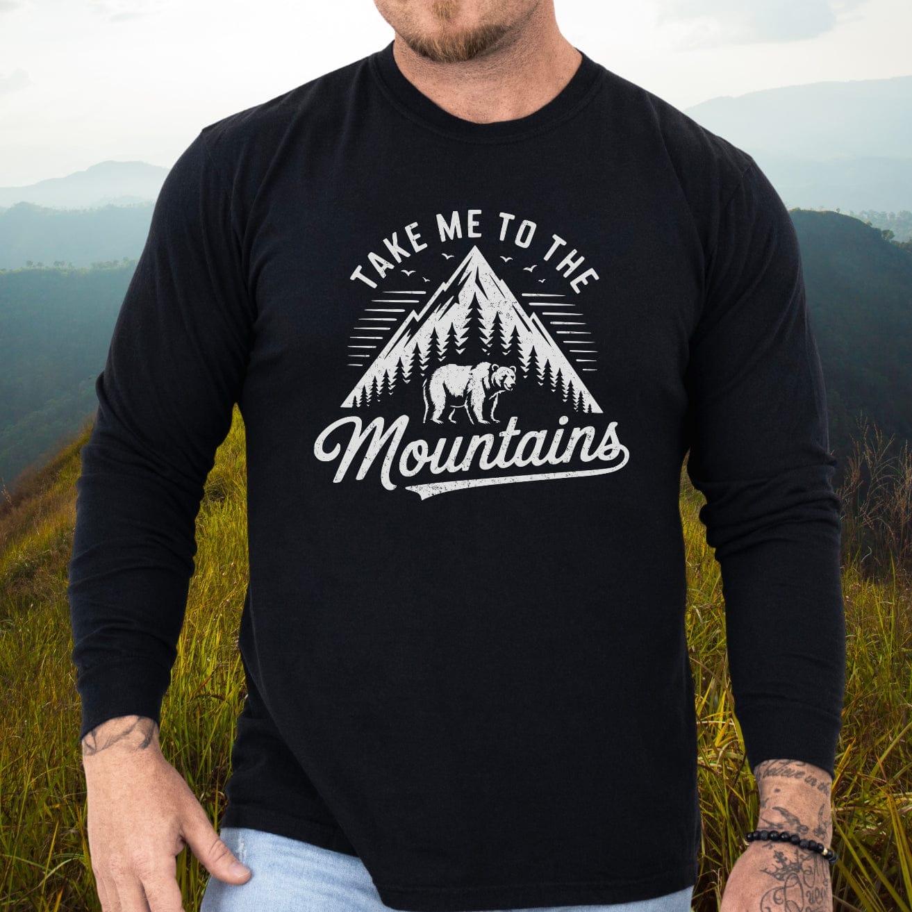 Person wearing a black Mountain Energy Comfort Colors Long-Sleeve Tee with mountain graphic, standing on a grassy hillside, showcasing tattoos and a beaded bracelet.