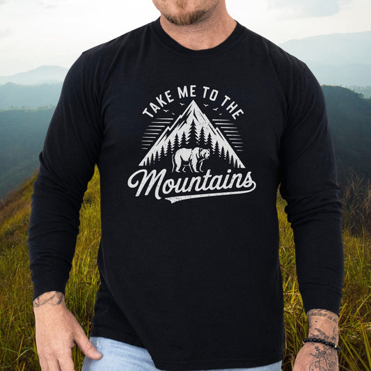 Person wearing a black Mountain Energy Comfort Colors Long-Sleeve Tee with mountain graphic, standing on a grassy hillside, showcasing tattoos and a beaded bracelet.