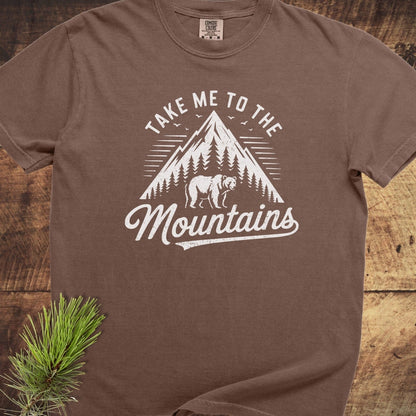 Take Me To The Mountains Comfort Colors 1717 Tee with mountain and bear design, displayed on wood, features durable, relaxed fit, 100% cotton, soft-washed fabric.