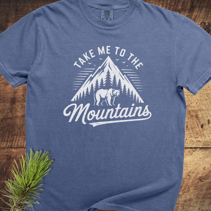 Take Me To The Mountains Comfort Colors 1717 Tee, featuring a mountain and bear graphic, laid flat with pine sprig, highlighting its relaxed fit and durability.