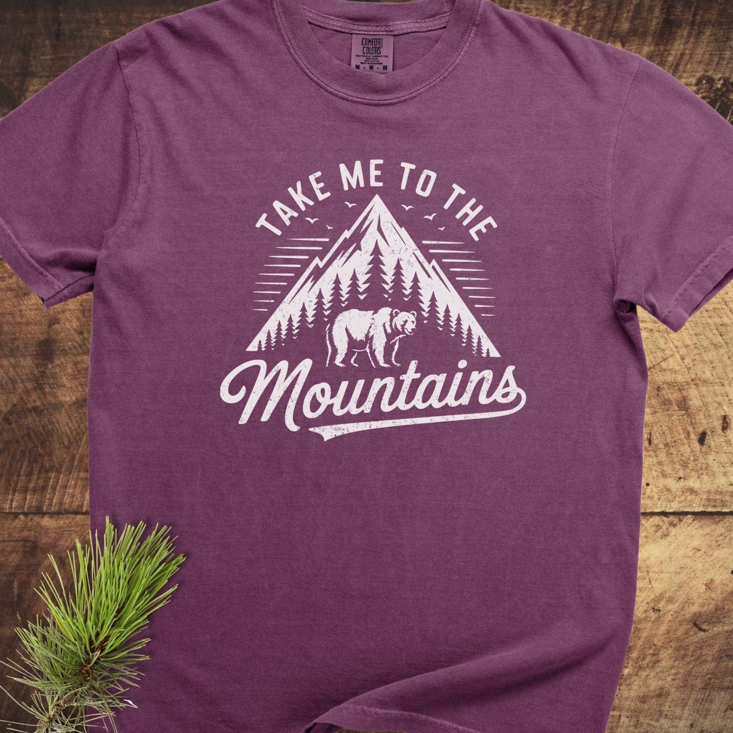 Take Me To The Mountains Comfort Colors 1717 Tee featuring a mountain peak graphic with trees and a bear, laid flat on a wooden surface.