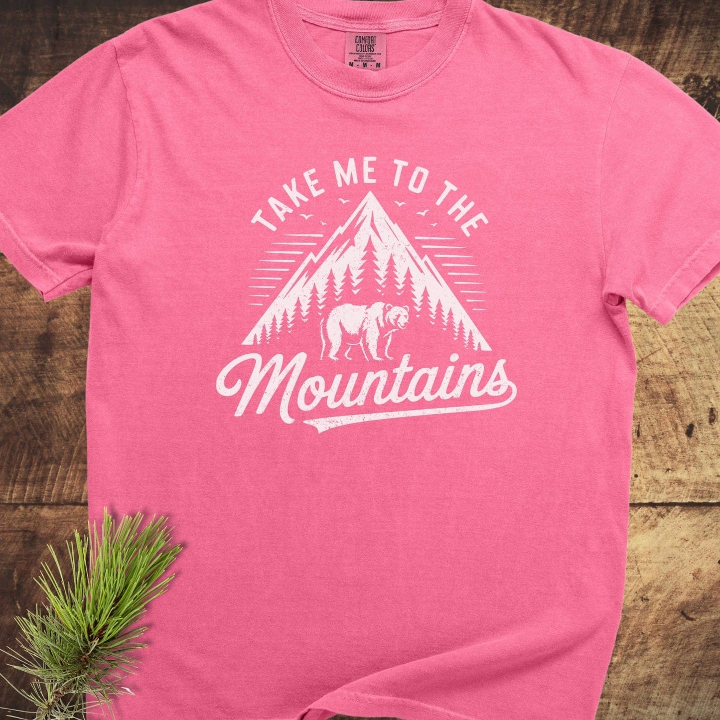 Take Me To The Mountains Comfort Colors 1717 Tee featuring a mountain graphic, pine trees, and bear silhouette, laid flat on wood with pine sprig accent.