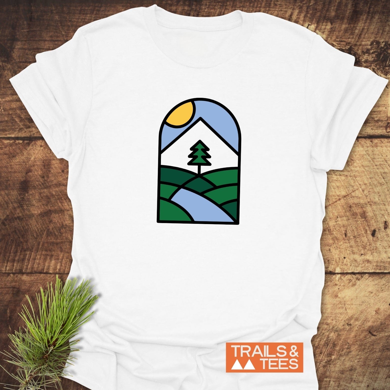 Minimalist Mountain T-Shirt featuring a stained-glass style nature scene with mountain, sun, and river, laid on wood with TRAILS & TEES logo visible.