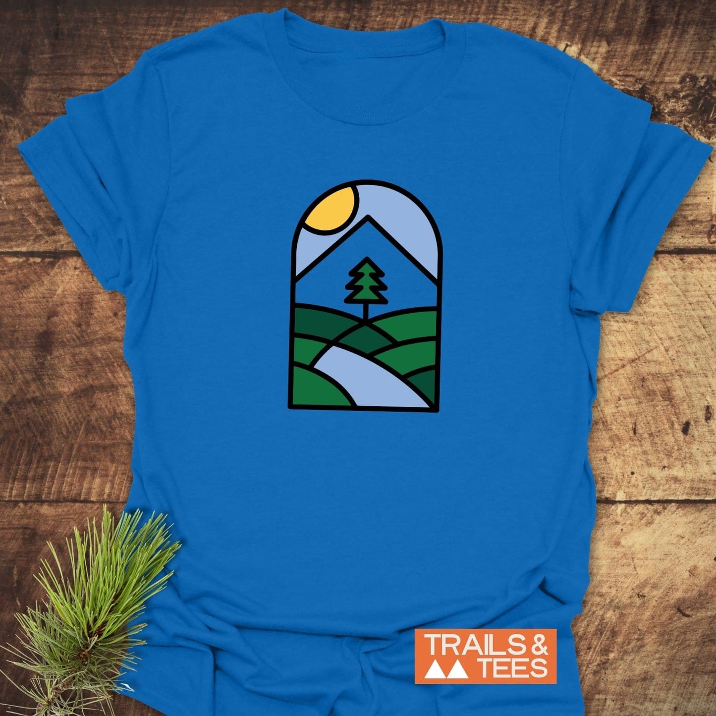 Minimalist Mountain T-Shirt featuring a geometric landscape graphic on bright blue fabric, styled with pine needles, showcases trail-tested comfort and durable DTF prints.