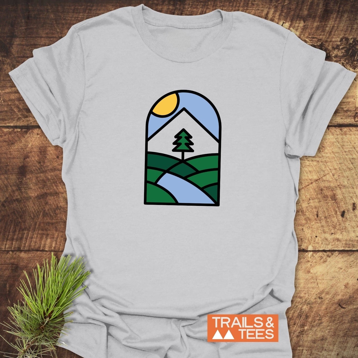 Minimalist Mountain T-Shirt featuring a nature graphic with hills and a sun, laid on wood with a pine sprig, branded TRAILS & TEES.