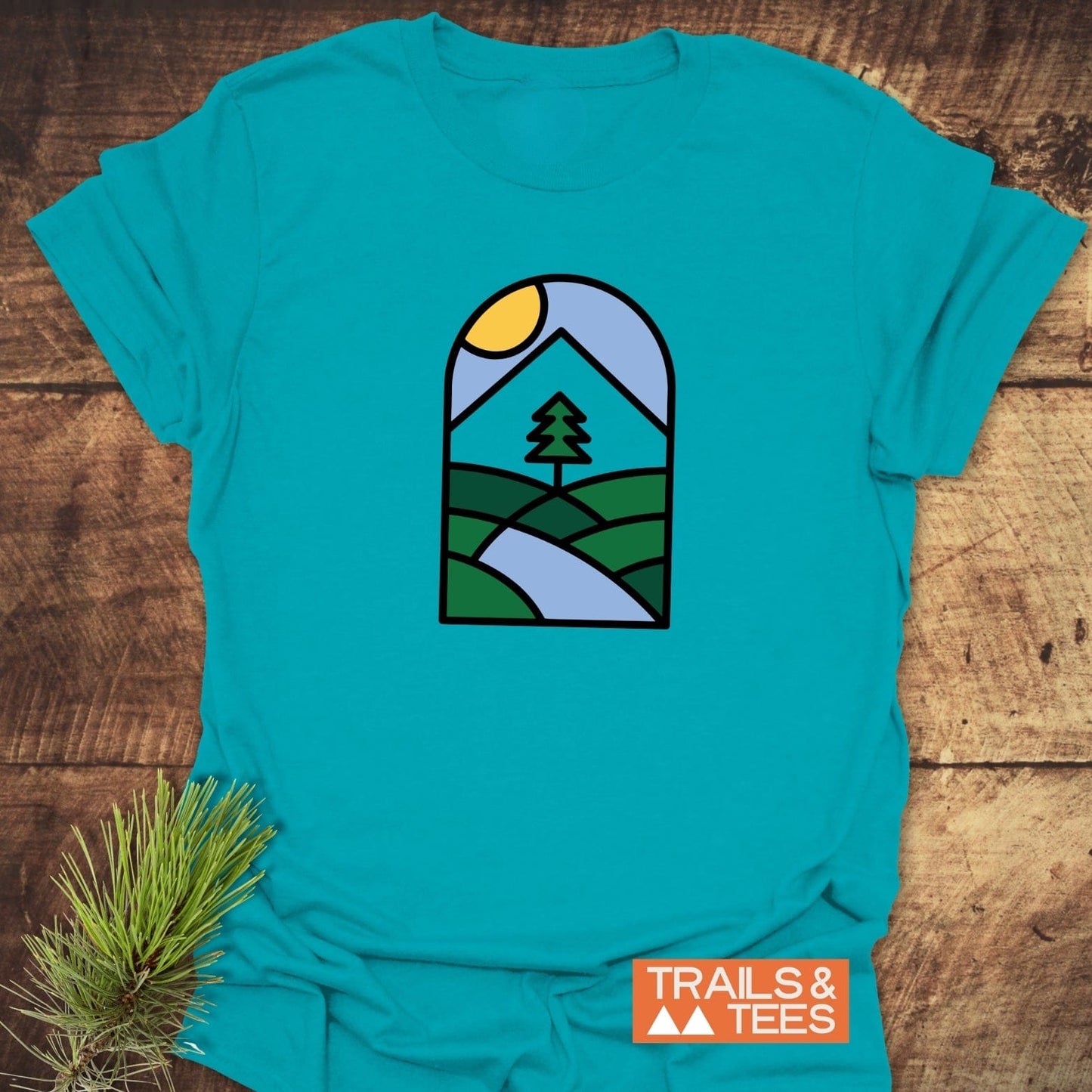 Minimalist Mountain T-Shirt featuring an outdoor scene graphic, laid on wood with pine sprig. Offers trail-tested comfort in soft, durable cotton.