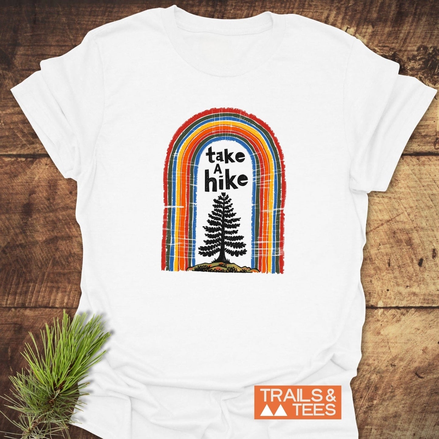 Take A Hike Rainbow T-Shirt on wooden surface, features multi-colored arch and conifer silhouette. Includes TRAILS & TEES logo, showcasing outdoor adventure spirit.