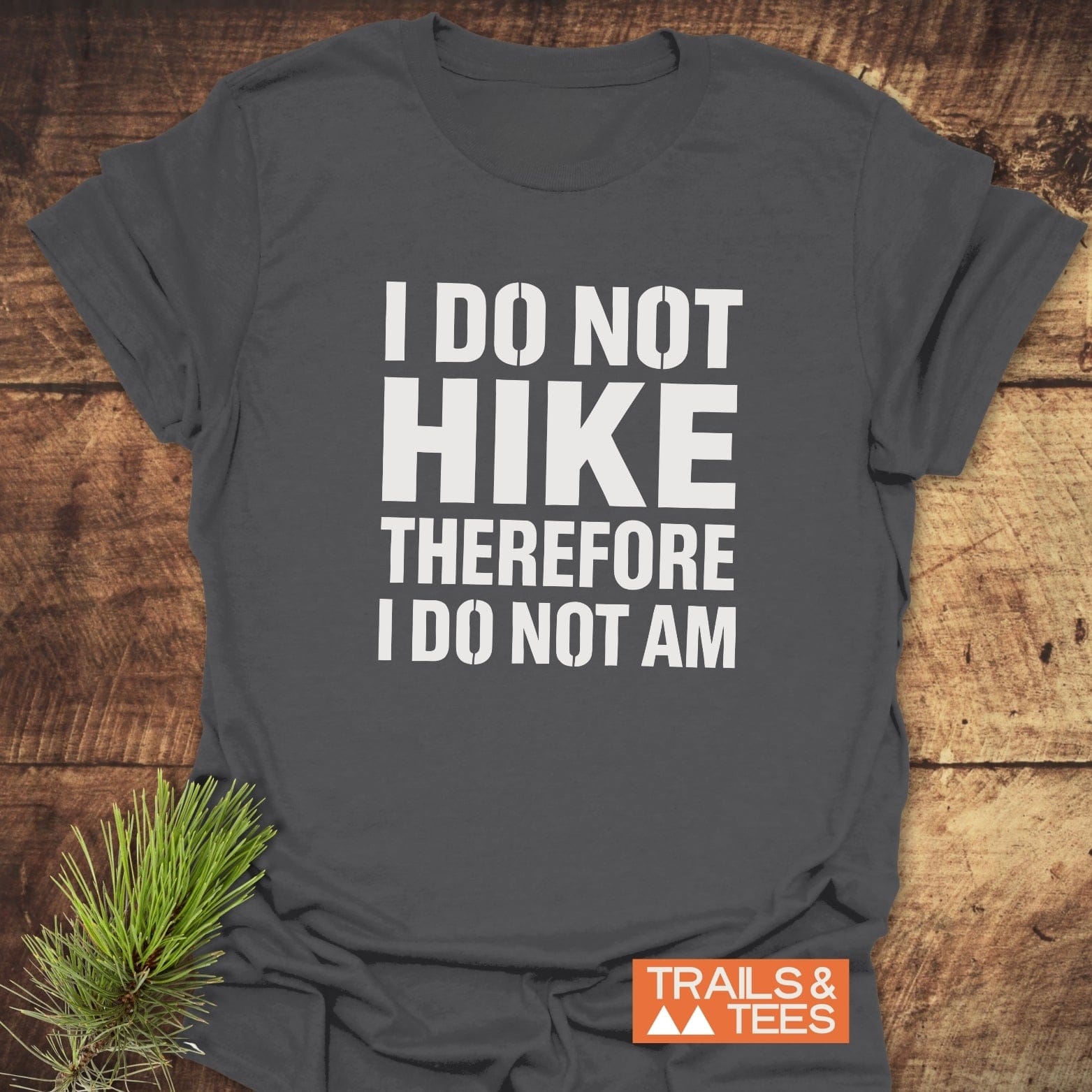 Charcoal-gray I Do Not Hike Funny T-Shirt with bold white lettering, TRAILS & TEES logo, and pine sprig on a wood-plank surface.