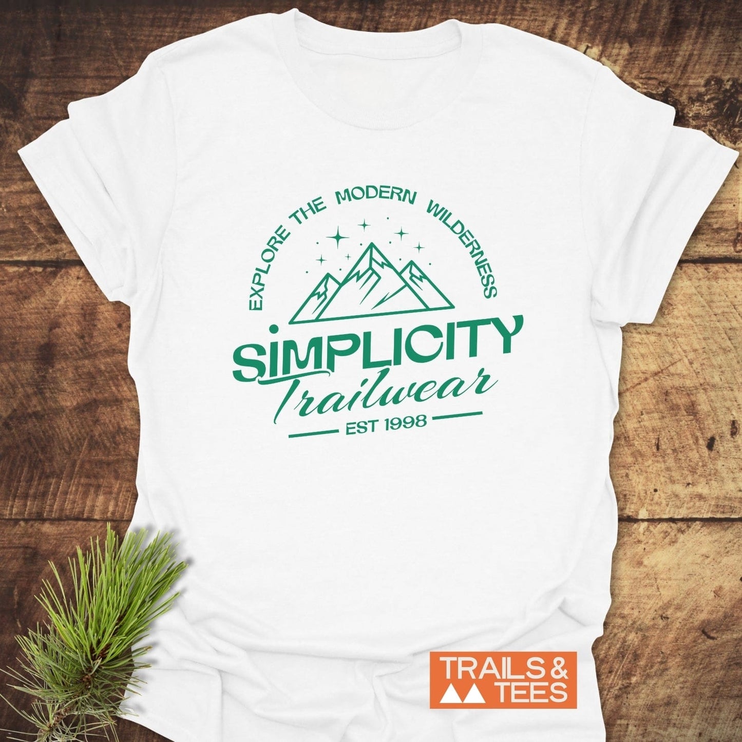 Simplicity Trailwear T-Shirt featuring a forest-green outdoor logo with text Explore the Modern Wilderness and EST 1998 on a wooden surface.