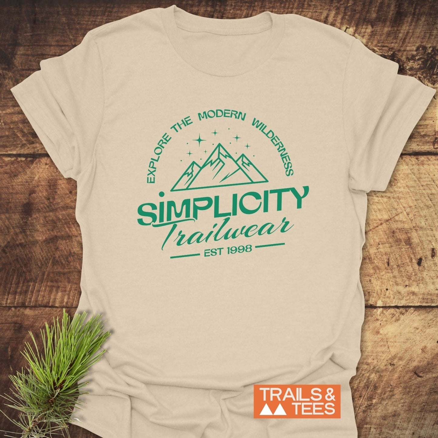 Simplicity Trailwear T-Shirt with green mountain logo and Explore the Modern Wilderness slogan, orange TRAILS & TEES badge, on rustic wooden surface.