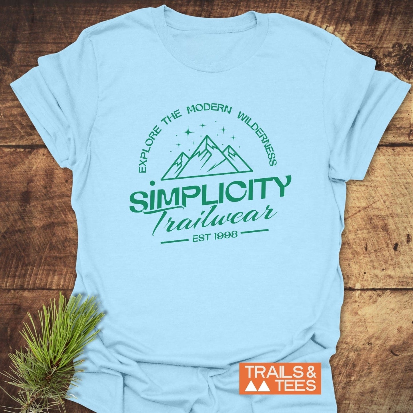 Simplicity Trailwear T-Shirt featuring a mountain graphic with stars, slogan, and logo. Laid on wood with a pine sprig beside.