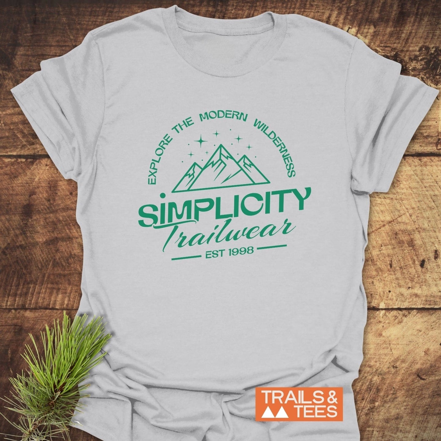 Simplicity Trailwear T-Shirt featuring a green mountain range logo with “Explore the Modern Wilderness” slogan, laid on wood with “TRAILS & TEES” badge visible.