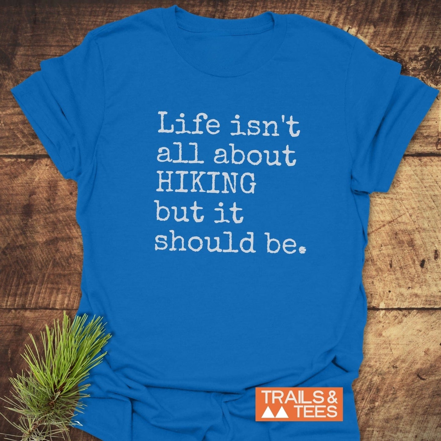 Bright blue “Life Isn't All About Hiking T-Shirt” with a white slogan, orange “TRAILS & TEES” logo, and pine branch accent on rustic wood surface.