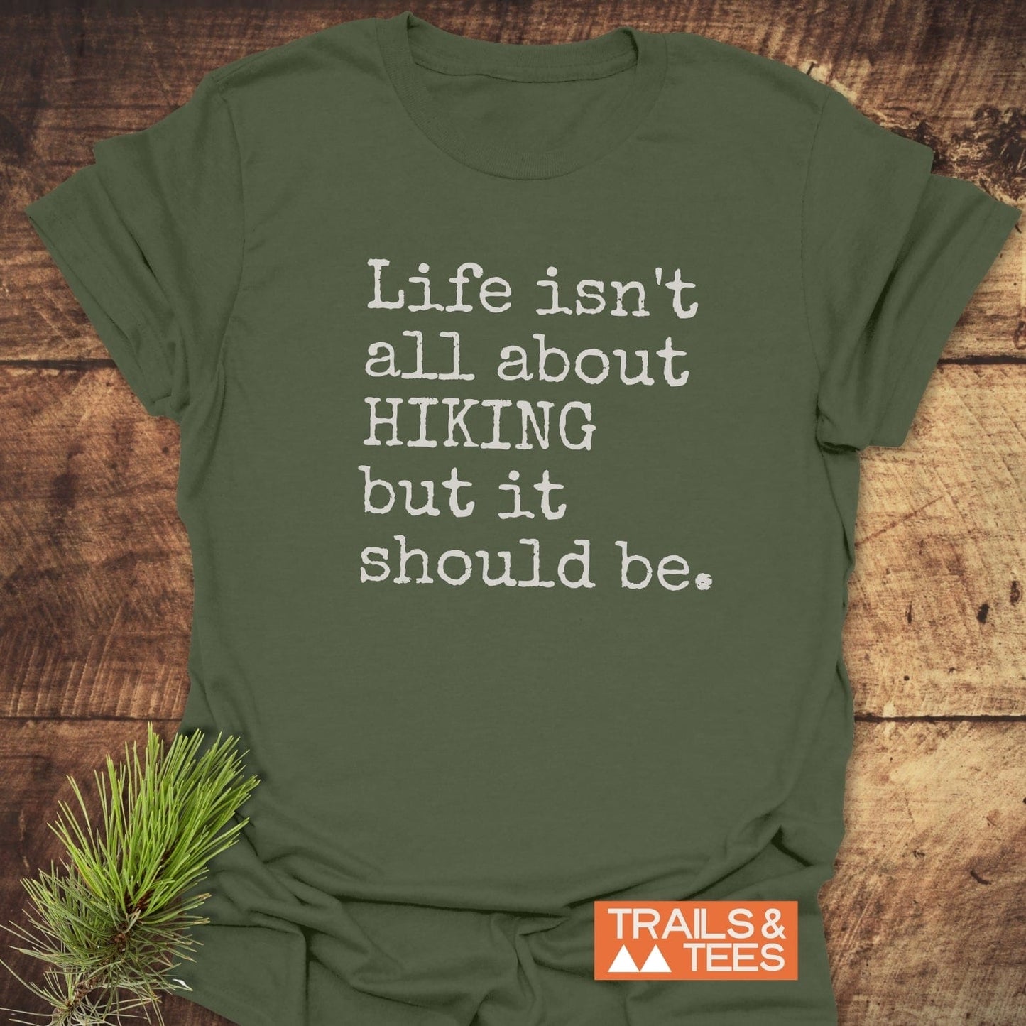 Life Isn't All About Hiking T-Shirt with white typewriter-style text, displayed on wooden background with pine sprigs, featuring TRAILS & TEES logo.