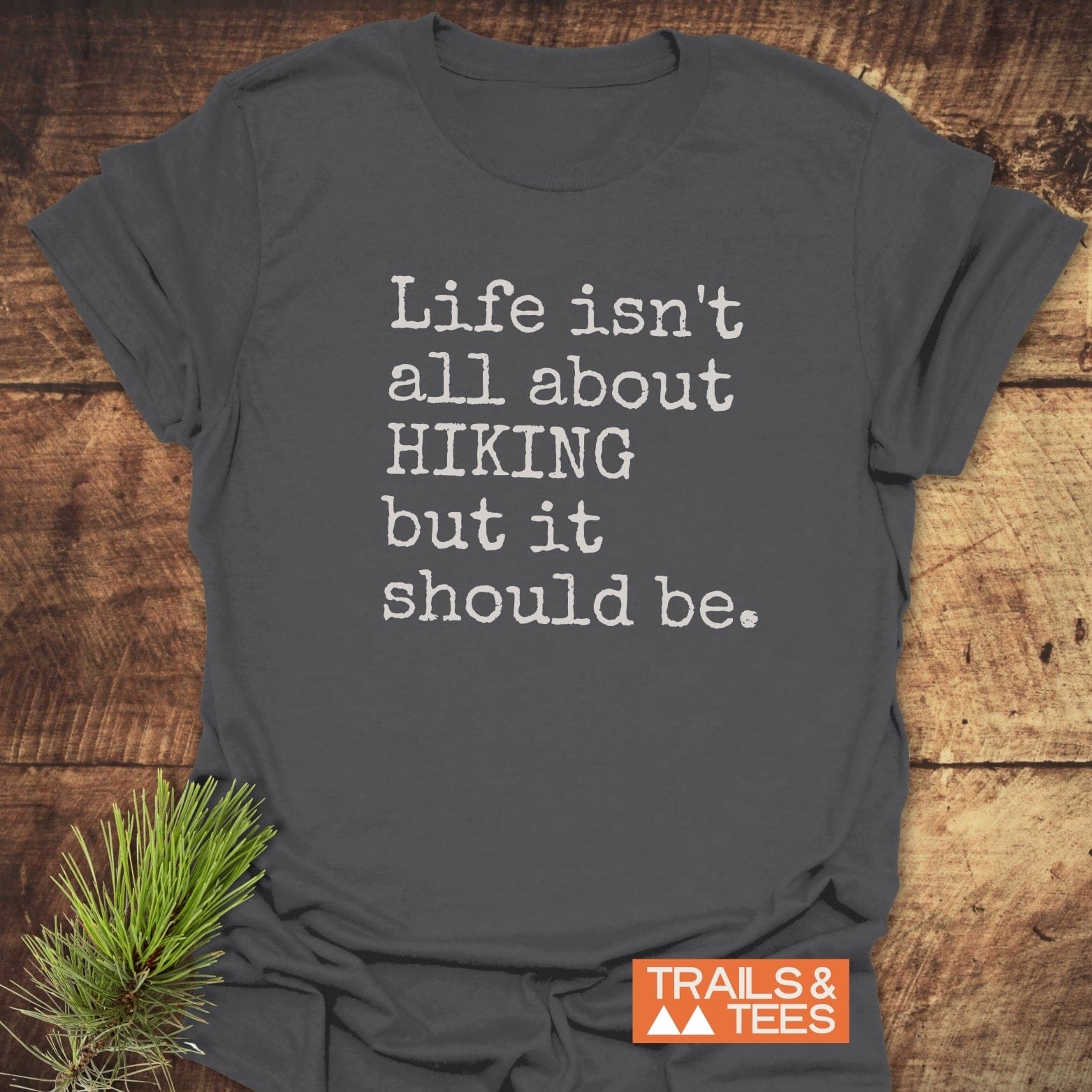 Life Isn't All About Hiking T-Shirt on wooden surface with slogan in distressed font, featuring Trails & Tees logo and pine needle sprig accent.
