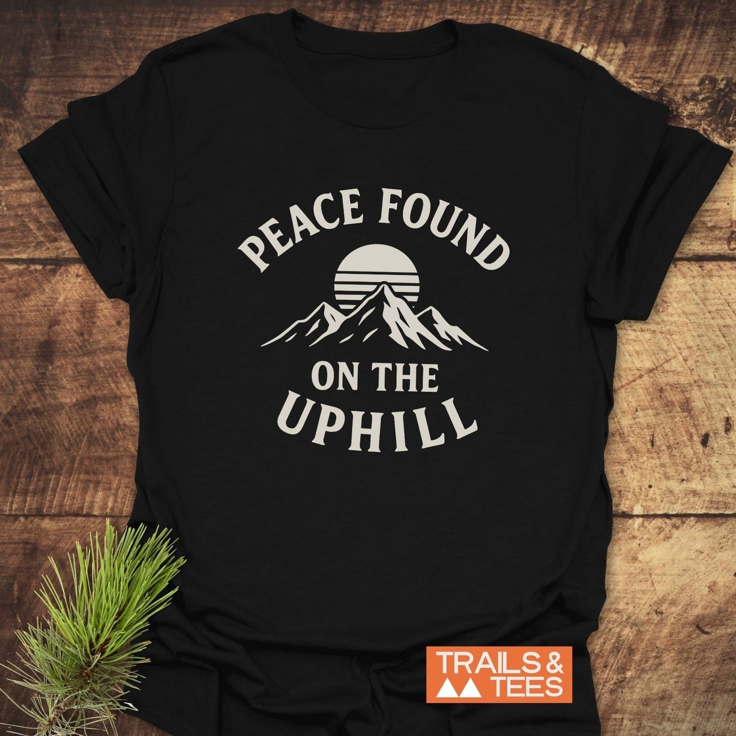 Peace Found T-Shirt with mountain graphic and slogan “PEACE FOUND ON THE UPHILL,” featuring Trails & Tees label, displayed on a wooden surface.