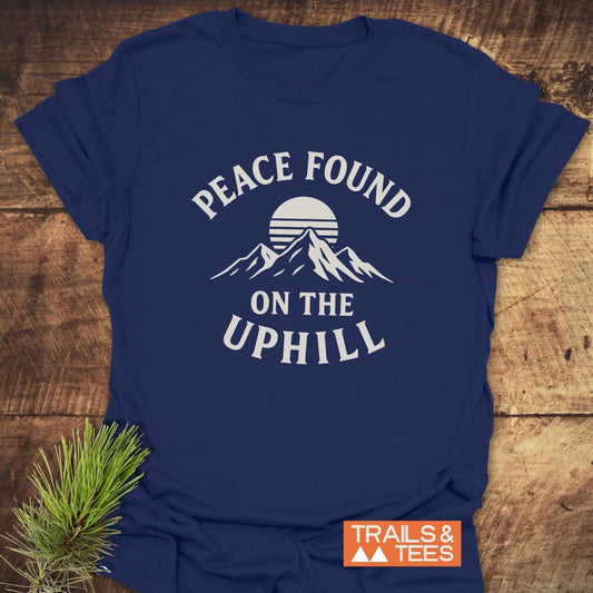 Peace Found T-Shirt with mountain and sun graphic, PEACE FOUND ON THE UPHILL, and TRAILS & TEES logo, displayed on a wooden surface.
