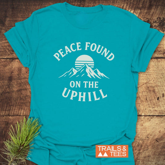 Peace Found T-Shirt featuring a mountain graphic and sun with “PEACE FOUND ON THE UPHILL” slogan, beside a small pine sprig on wood.