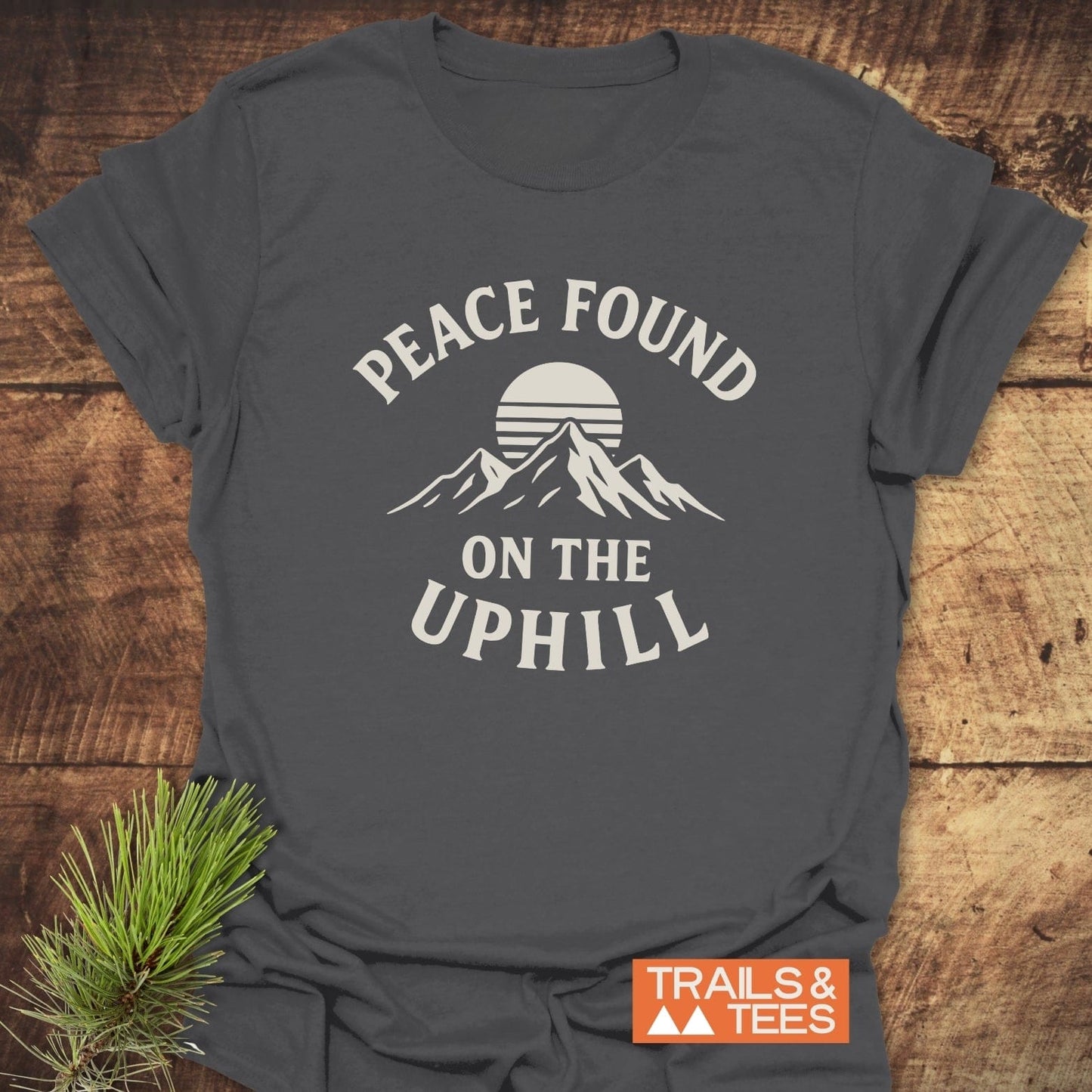 Peace Found T-Shirt featuring a stylized sun and mountains graphic with the phrases PEACE FOUND and ON THE UPHILL. Branded by Trails & Tees.