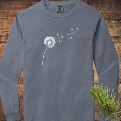 Dandelion Comfort Colors Long-Sleeve Tee featuring a stylized dandelion print, laid flat on wood, with a pine needle sprig in the corner.