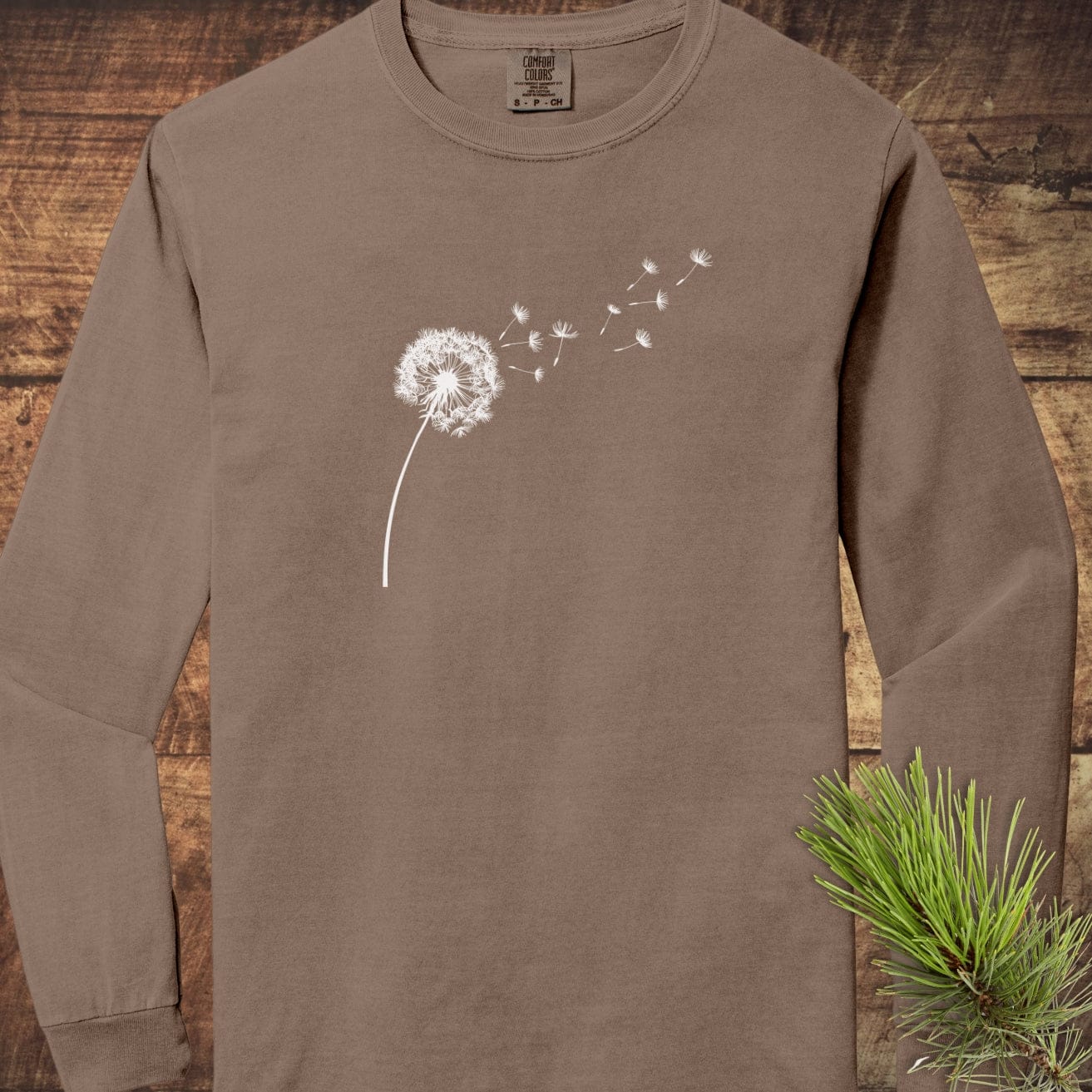 Dandelion Comfort Colors Long-Sleeve Tee with white dandelion design on taupe, laid on wood. Features relaxed fit and soft, 100% ring-spun cotton fabric.