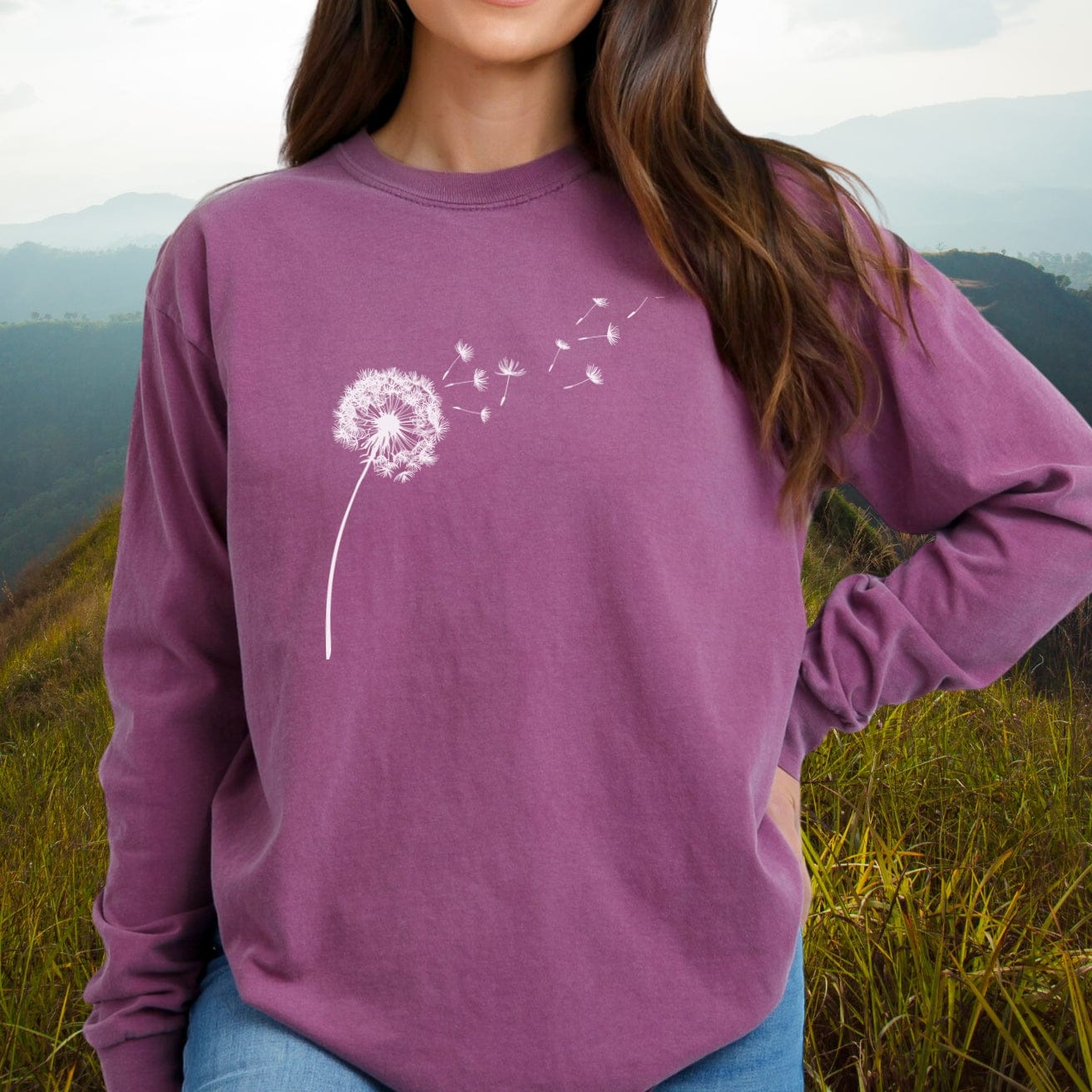Person wearing Dandelion Comfort Colors Long-Sleeve Tee with white dandelion graphic, relaxed fit, on a grassy hillside. Made from soft, 100% ring-spun cotton.