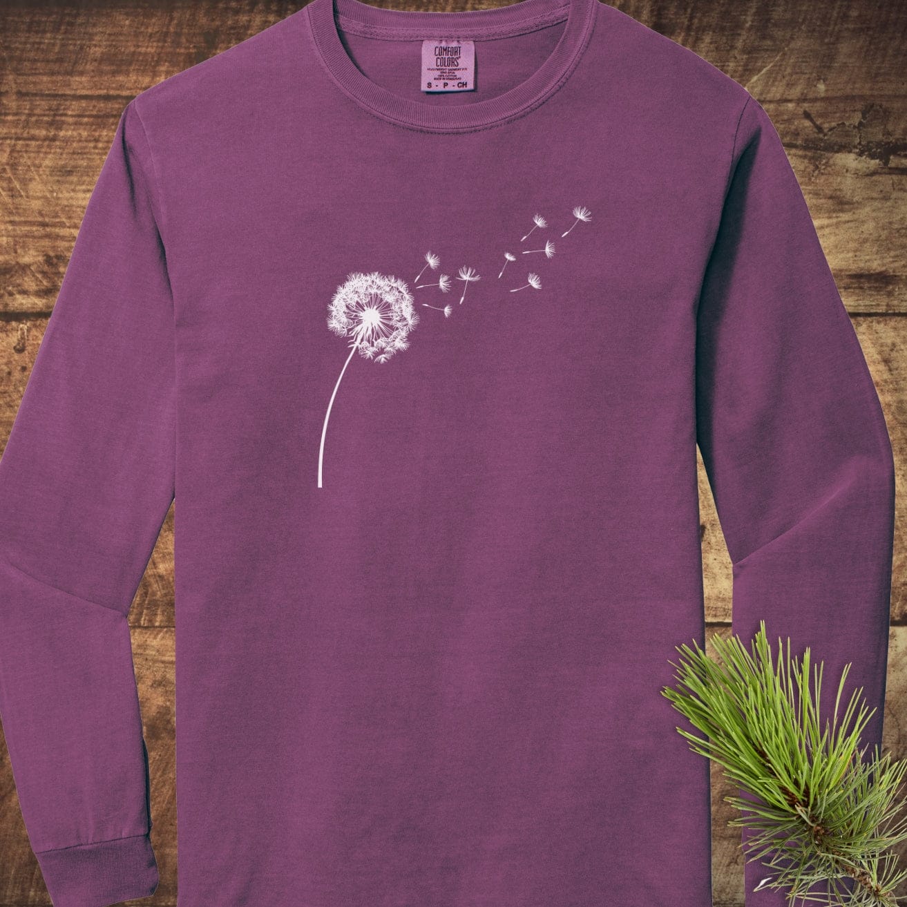 Dandelion Comfort Colors Long-Sleeve Tee featuring a white stylized dandelion design on soft, garment-dyed cotton, laid on wooden surface with pine sprig.