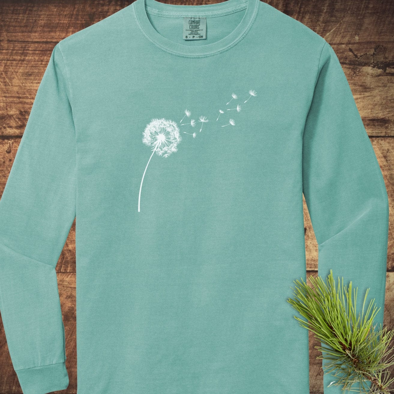 Dandelion Comfort Colors Long-Sleeve Tee in teal, featuring a white dandelion print on chest, laid flat on a wooden surface with a pine sprig.