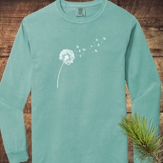 Dandelion Comfort Colors Long-Sleeve Tee in teal, featuring a white dandelion print on chest, laid flat on a wooden surface with a pine sprig.