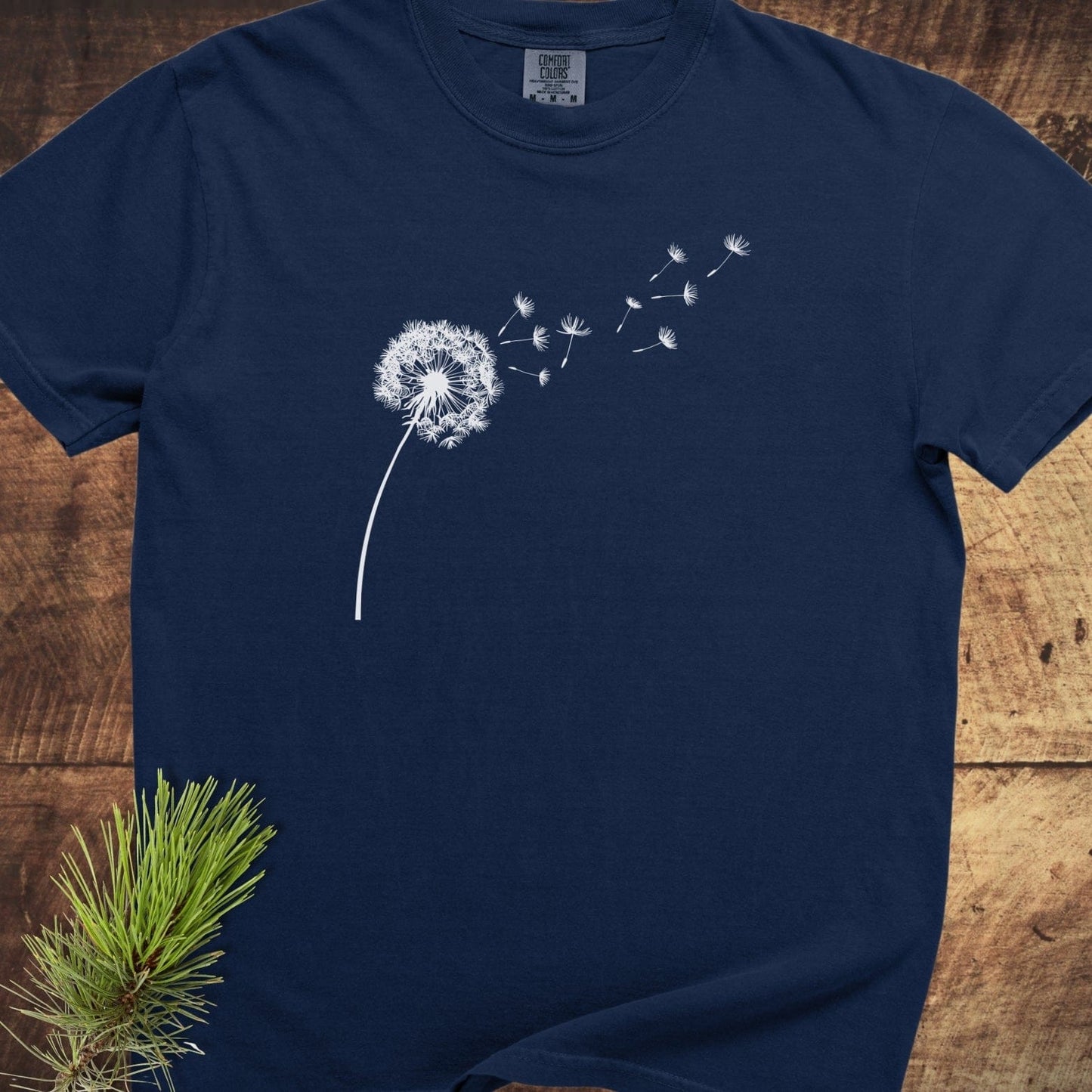 Dandelion Comfort Colors 1717 Tee, navy-blue, features a white dandelion illustration, laid flat on wood, with durable double-needle stitching and relaxed fit.