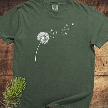 Forest-green Dandelion Comfort Colors 1717 Tee with white dandelion graphic, laid on wood surface, featuring a pine sprig; showcasing its relaxed, durable, garment-dyed design.