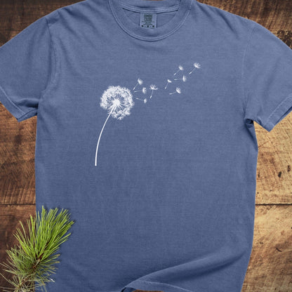 Dandelion Comfort Colors 1717 Tee in heather-blue with a white dandelion graphic, on a wooden surface, accompanied by a pine sprig.